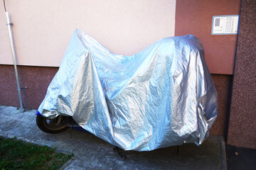 The motorcycle is parked by the sidewalk on the road and is covered with a protective tarpaulin