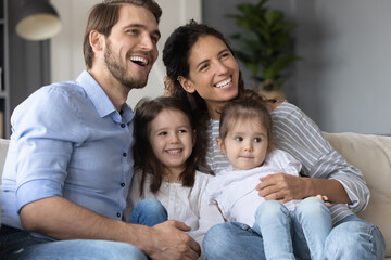 Smiling young married couple relaxing on sofa with adorable little children daughters, enjoying watching TV program, kids show or cartoons together at home, happy family hobby weekend pastime.