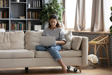 Obraz premium Full length smiling young attractive barefoot woman in eyeglasses sitting on sofa with computer on laps, enjoying working remotely from home, using laptop applications, shopping online or web surfing.