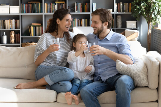 Happy young couple parents teaching little preschool daughter drinking clear water every day. Smiling healthy family holding glasses with pure aqua, enjoying morning daily healthcare habit at home.