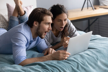 Surprised young family couple looking at computer screen, lying on bed. Shocked millennial man and woman reading email with unbelievable news, feeling astonished together in bedroom, omg reaction. - obrazy, fototapety, plakaty