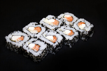 Sushi roll with cheese and salmon sprinkled with black sesame seeds on a black background.