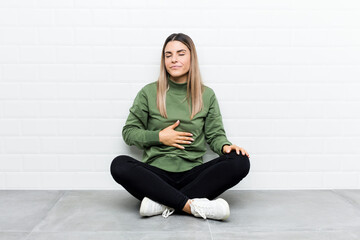 Young caucasian woman sitting on the floor touches tummy, smiles gently, eating and satisfaction concept.