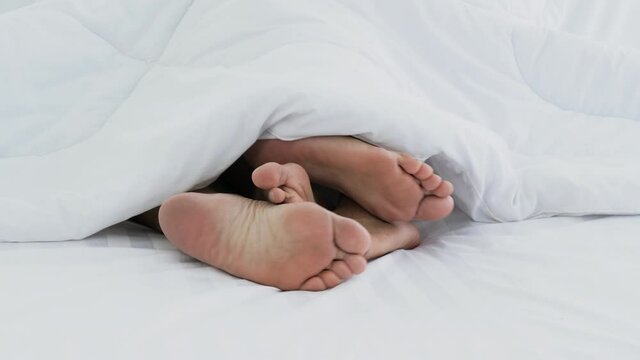 Close up of young man and loving couple hug and play their feet each other under blanket while wake up on bed in morning.