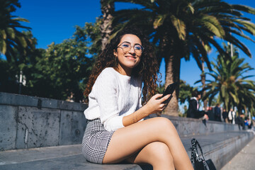 Positive female sitting on stone bench with smartphone