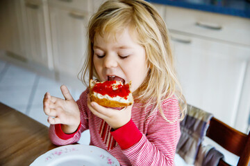Cute funny toddler girl eats sweet bun for breakfast. Happy child eating bread roll with strawberry...