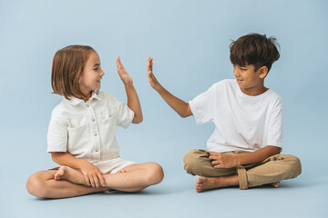 Little boy and girl of roughly the same age sitting on the floor cross-legged. Over blue background. Looking at each other, giving high five.