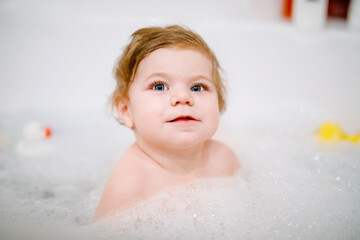 Cute adorable baby girl taking foamy bath in bathtub. Toddler playing with bath rubber toys. Beautiful child having fun with colorful gum toys and foam bubbles