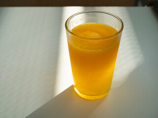 Glass of freshly squeezed orange juice on white table illuminated by a ray of sunshine, liquid orange extract rich in vitamin c
