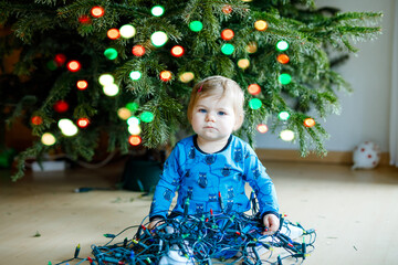 Cute baby girl taking down holiday decorations from Christmas tree. child holding light garland. Family after celebration remove and dispose tree