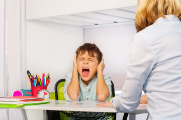 Autistic boy scream and close ears in pain during ABA development therapy sitting by the table with...