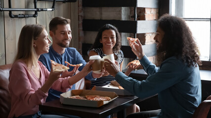 Happy diverse friends eating pizza and having fun in cafe, sitting at table together, overjoyed young men and women laughing at funny joke, holding Italian junk food slices and coffee cups