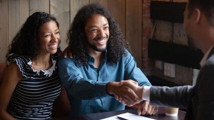 Smiling African American man shaking realtor manager advisor hand at meeting, family making deal, happy young wife and husband planning wedding, purchasing new apartment or taking mortgage