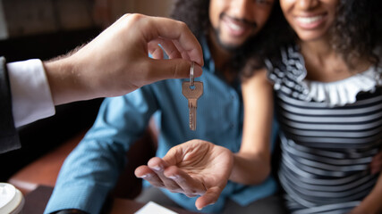Close up happy young African American couple receiving keys from realtor, real estate agent, overjoyed family husband and wife purchasing first apartment, buying new house, making deal at meeting