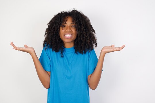 Clueless Young african woman with curly hair wearing casual blue shirt over white background, shrugs shoulders with hesitation, faces doubtful situation, spreads palms, Hard decision