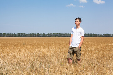 Naklejka na ściany i meble Young Caucasian man wearing shorts and white t-shirt standing in wheat field and looking far away, copy space
