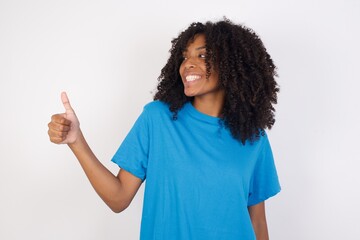 Young african woman with curly hair wearing casual blue shirt Looking proud, smiling doing thumbs up gesture to the side. Good job!