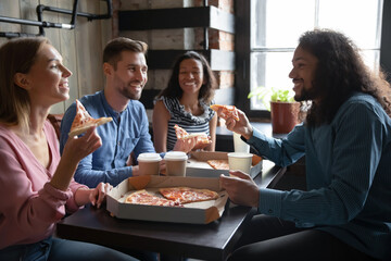 Overjoyed smiling diverse friends enjoying pizza in cafe on weekend together, excited happy young people sitting at table, laughing at joke, drinking coffee or tea, holding slices and cups