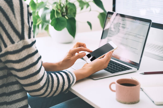 Woman Using Smartphone While Working On Laptop In Home Office.