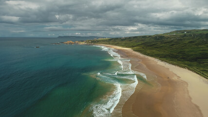 Ireland aerial view: beach ocean bay water crashing. People on shore walk and playing with dogs. Meadows in horizon. Picturesque Northern beauty