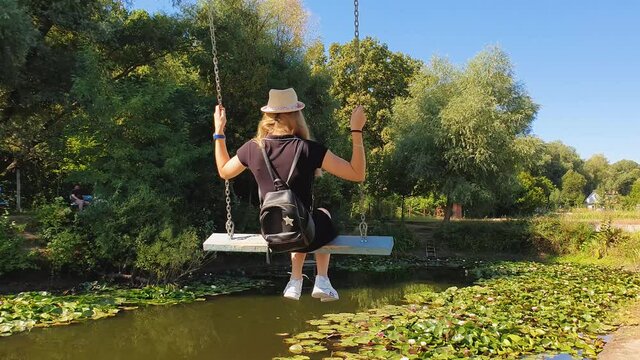 Carefree young woman sway on a swing over a pond with blooming waterlily, lotus flowers. Vacation travel, natural background at the lake with a beautiful fit girl happy swinging, at Bardar, Moldova.