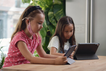 Two multi racial beautiful school age 9s girls sit at desk with portable computer. Using modern device ignoring each other, studying distantly by wireless gadget, new generation tablet overuse concept
