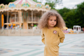 A beautiful little curly girl in an amusement park near the carousel in an ethnic yellow dress.
