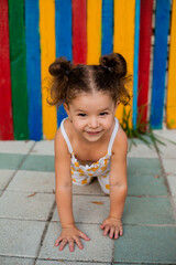 Fototapeta na wymiar Little girl crawls on the asphalt against the background of a wooden multi-colored fence