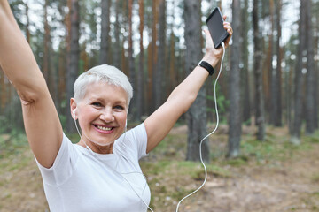 Energetic cheerful retired female with fit slim body posing outdoors in earphones, raising hands,...