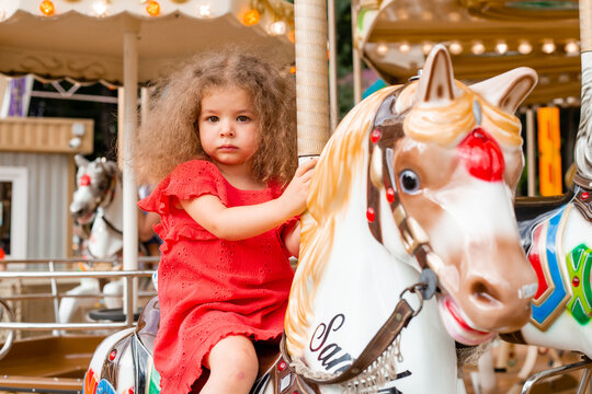 A little curly girl in a red dress sits on a horse in an amusement park on a carousel