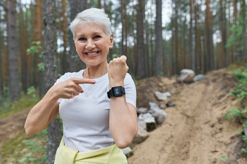 Happy fit retired female in activewear smiling broadly pointing at display of wrist smart watch, proud of calories burnt after physical activity. Mature woman using fitness tracker during morning run