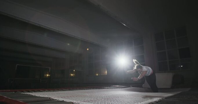 Girl gymnast doing coup exercise on his hands jumping on a trampoline. Active exercise on a trampoline in a sports form in a dark gym with single bright spotlight.