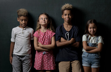 Four adorable cute multinational children school age girls boys looking at camera pose lean at dark grey wall studio background. Alpha generation beautiful different ethnicity kids portrait concept