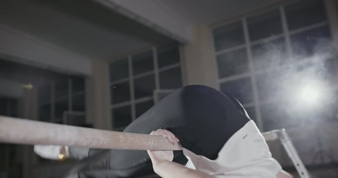 Girl gymnast does the exercise coup on the bars in the gym, focused exercise in a dark gym with single bright spotlight. Night gymnastics training of a girl in a sports hall at night.