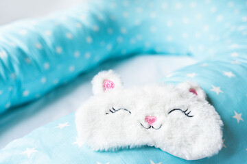 Fototapeta na wymiar Light blue supporting pillow with stars on white sheet in bed for comfortable sleep of pregnant woman. Funny sleeping mask in form of bunny,cat with eyes,ears,mouth. Accessories for expectant mothers