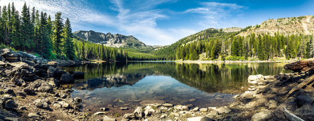 Panorama of a mountain lake with rocks in the foreground and trees in the far shore with mountains...