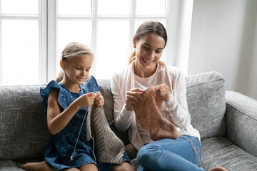Smiling cute smart little kid girl resting on sofa with older sister or biracial nanny, involved in knitting warm clothes at home. Happy skilled small daughter enjoying hobby activity with mother.