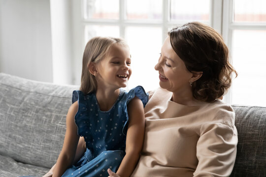 Smiling older senior woman sitting on cozy sofa with energetic little granddaughter, communicating at home. Happy small kid girl enjoying funny conversation, laughing at joke with caring granny.