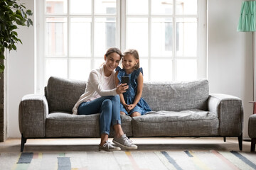 Full length smiling young biracial woman sitting on comfortable sofa with younger sister or small pretty daughter, posing for selfie photo on smartphone or enjoying using mobile application at home.