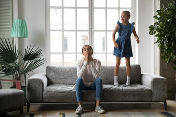 Full length energetic crazy little kid girl jumping on sofa near exhausted young biracial mother or babysitter. Stressed millennial woman feeling tired of loud naughty small child daughter at home. - obrazy, fototapety, plakaty