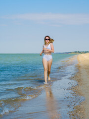 Fototapeta premium A young girl runs along the sandy sea beach along the surf. Splashes of sea water. The girl is wearing a white swimsuit and sunglasses. Freedom and carelessness. Outdoor fitness. Sunny day. Copy space