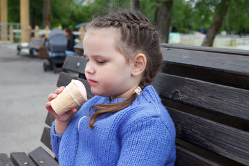 child girl in a blue knitted sweater eats ice cream sitting on a bench