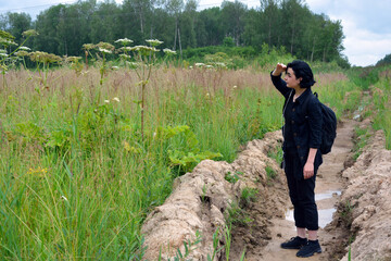 Fototapeta na wymiar A girl with a backpack on her shoulders stands in a ditch in a field and looks into the distance, raising her hand to her forehead.