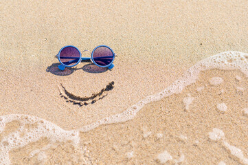 Fototapeta na wymiar A symbol of good mood and positive seaside relaxation. The smile is painted on the seaside next to the sunglasses. Sea wave on a sandy shore