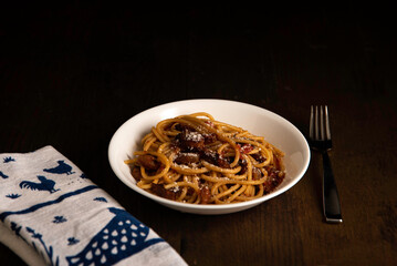 Prepared pasta and sauce in a white bowl white and blue cloth and metal fork on a dark wood table with moody lighting.