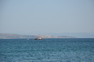 A small fishing boat and a small dinghy in the immense Aegean Sea. A background with a hazy view of distant hills and residential areas. The hills on the opposite shore appear in silhouette. 