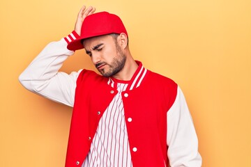 Handsome sporty man with beard wearing baseball jacket and cap over yellow background surprised with hand on head for mistake, remember error. Forgot, bad memory concept.