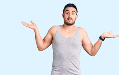 Fototapeta premium Young handsome man wearing swimwear and sleeveless t-shirt clueless and confused expression with arms and hands raised. doubt concept.
