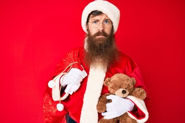 Handsome young red head man with long beard wearing santa claus costume holding teddy bear relaxed with serious expression on face. simple and natural looking at the camera.