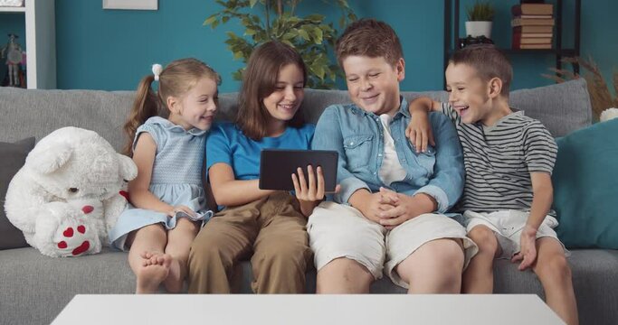 Group of four happy kids sitting together on grey couch and using one digital tablet. Smiling little people spending free time with fun at home. Concept of modern technology.